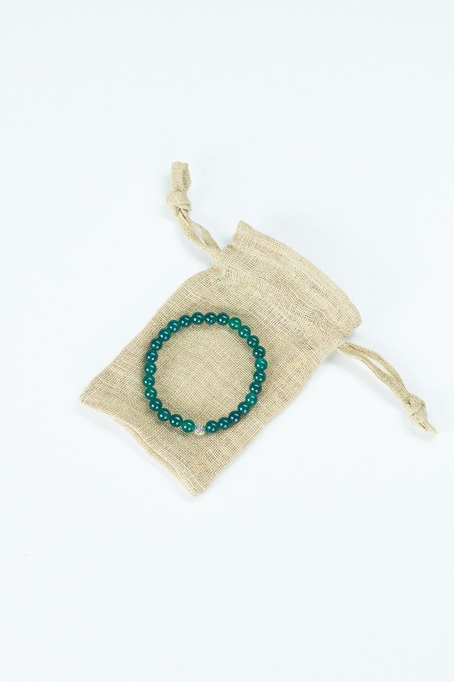 A beaded bracelet made of a green stone is positioned atop a small bag made out of recycled materials. The bag is white in color and slightly larger than the bracelet.