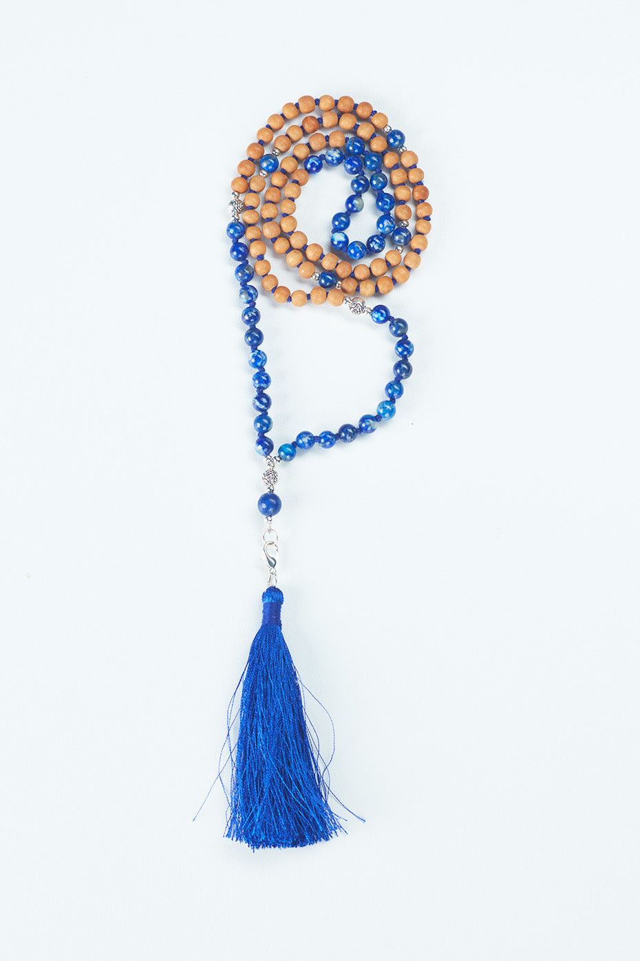 A beaded necklace that is in the center of the image, made of Sandalwood and a dark blue stone, with a dark blue tassel at the bottom