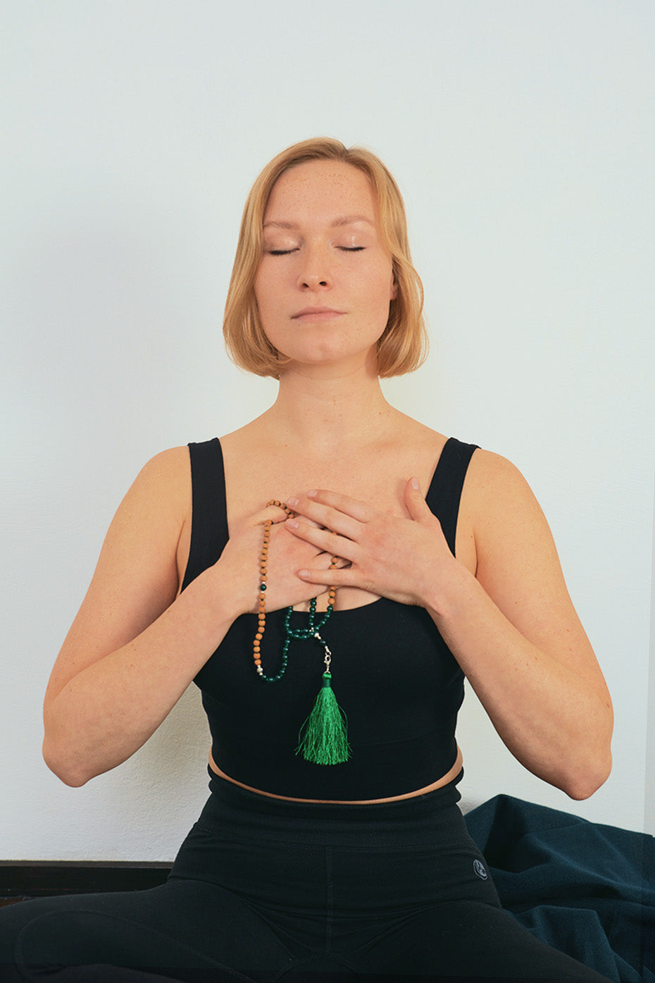  A woman seated with crossed legs, holding her hands to her chest with a beaded necklace held between her hands. 