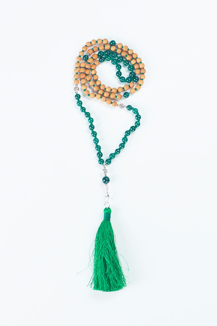  A beaded necklace that is in the center of the image, made of Sandalwood and a green stone, with a green tassel at the bottom.